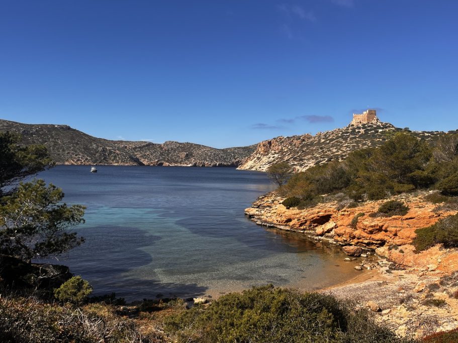 Vista de una bahía con agua clara y montañas al fondo bajo un cielo azul.