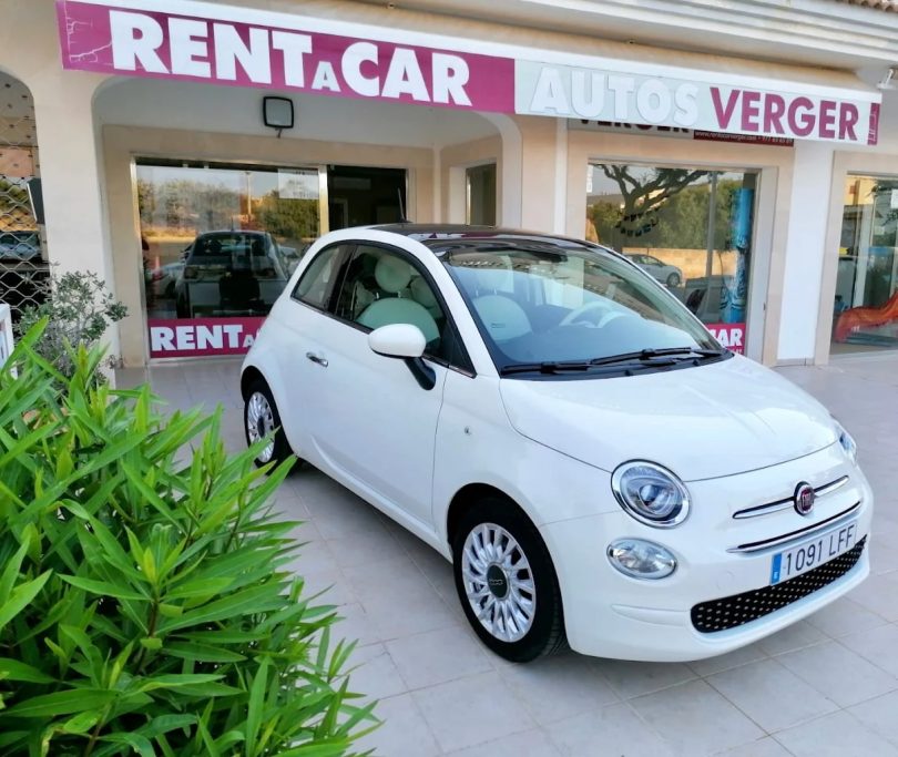 Coche blanco Fiat 500 estacionado frente a una oficina de alquiler de autos.