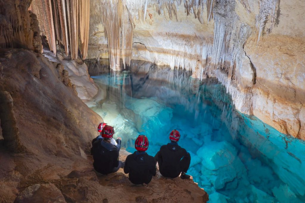 Tres personas con cascos sentadas junto a un lago subterráneo iluminado.